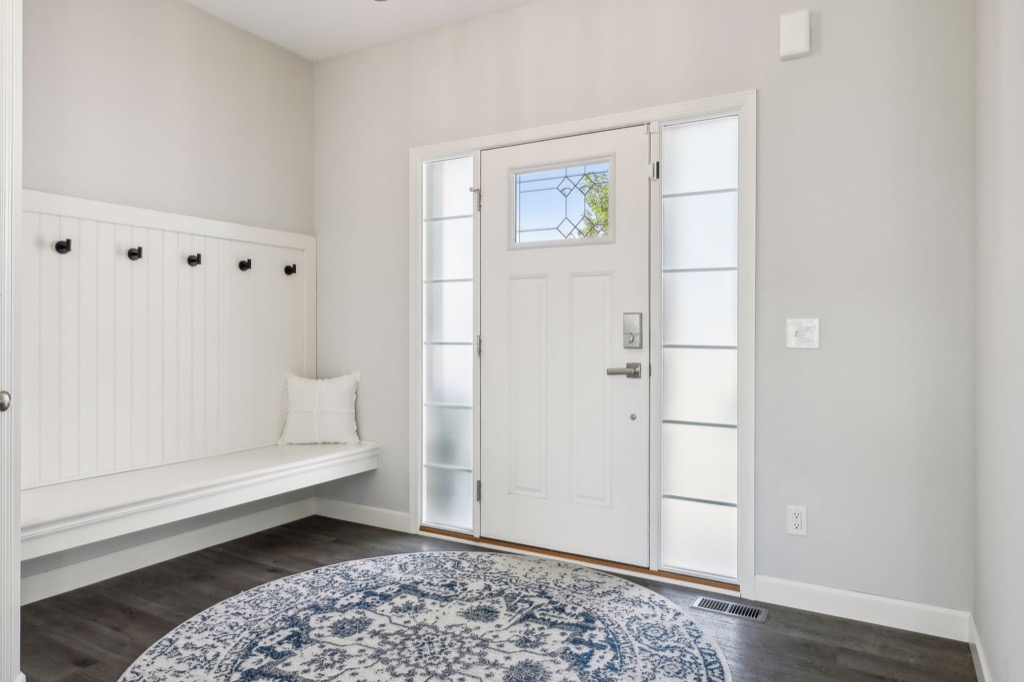 A bright, modern entryway on Fairchild Way with a white front door, frosted side windows, a patterned blue and white rug, and a built-in white bench with black hooks and a white pillow on dark wood floors—perfect for PHOTOS.