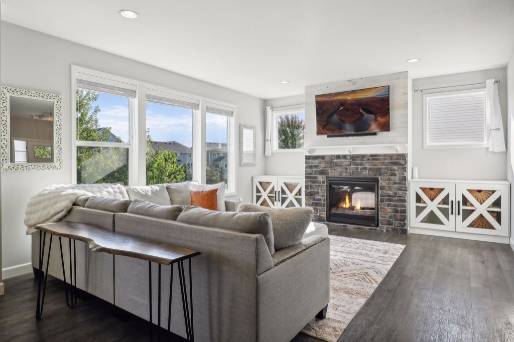 Modern living room on Fairchild Way with large windows, a grey sectional sofa, wooden console table, stone fireplace with TV above, and white cabinets—all in neutral tones and flooded with natural light. See PHOTOS for more details.