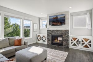 Bright living room on Fairchild Way with large windows, a gray sectional sofa with orange accent pillows, a patterned rug, stone fireplace, TV above the mantel, and built-in white cabinets. See PHOTOS for details.