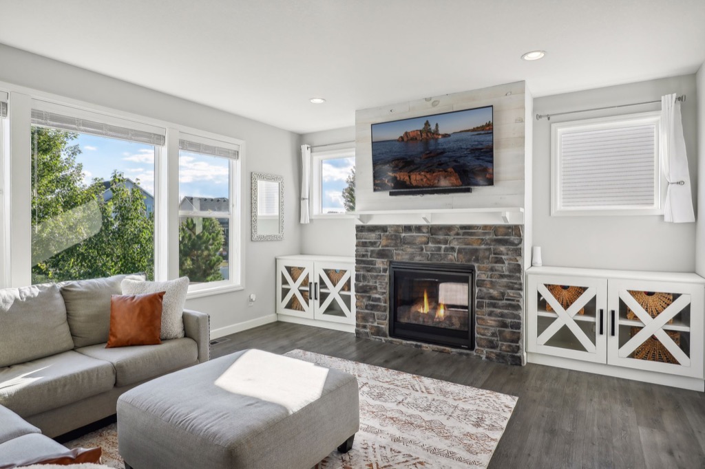 Bright living room on Fairchild Way with large windows, a gray sectional sofa with orange accent pillows, a patterned rug, stone fireplace, TV above the mantel, and built-in white cabinets. See PHOTOS for details.