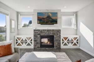 A modern living room at 15668 Fairchild Way features a stone fireplace, a TV above it displaying mountain PHOTOS, white cabinets on each side, gray flooring, and large windows bathing the space in natural light.