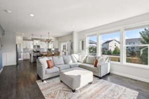Bright, modern open-concept living room and kitchen at 15668 Fairchild Way with a large sectional sofa, ottoman, patterned rug, dining table, and white cabinets. Large windows overlook a suburban neighborhood—see PHOTOS for more details.