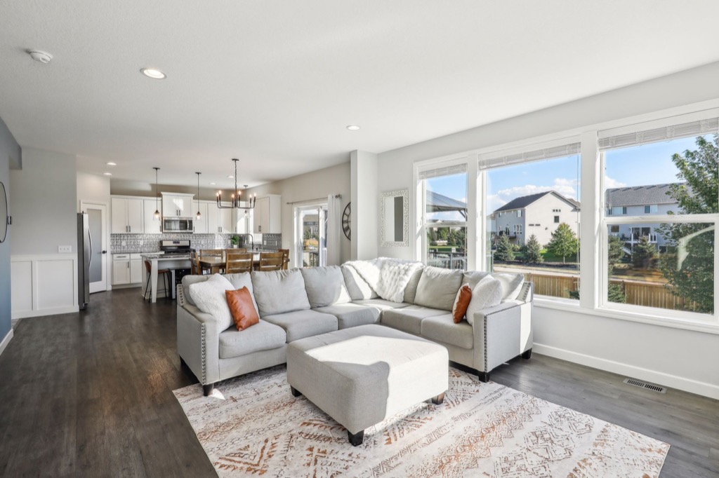 Bright, modern open-concept living room and kitchen at 15668 Fairchild Way with a large sectional sofa, ottoman, patterned rug, dining table, and white cabinets. Large windows overlook a suburban neighborhood—see PHOTOS for more details.