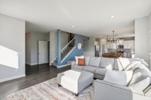 A bright, modern living room on Fairchild Way features a light gray sectional sofa, ottoman, and orange accent pillow. The open layout flows to a dining area and white kitchen with pendant lights—see PHOTOS for details.