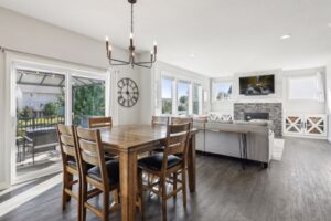 Bright, open-plan dining and living room on Fairchild Way with wood floors, wooden dining table and chairs, gray sofa, stone fireplace with TV above, large windows, and glass doors leading to an outdoor patio. See PHOTOS for details.