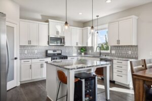 Modern kitchen at 15668 Fairchild Way with white cabinets, stainless steel appliances, a gray tile backsplash, granite countertops, a center island with two stools and a wine fridge, pendant lights, and a dining table with wooden chairs. See PHOTOS for details.