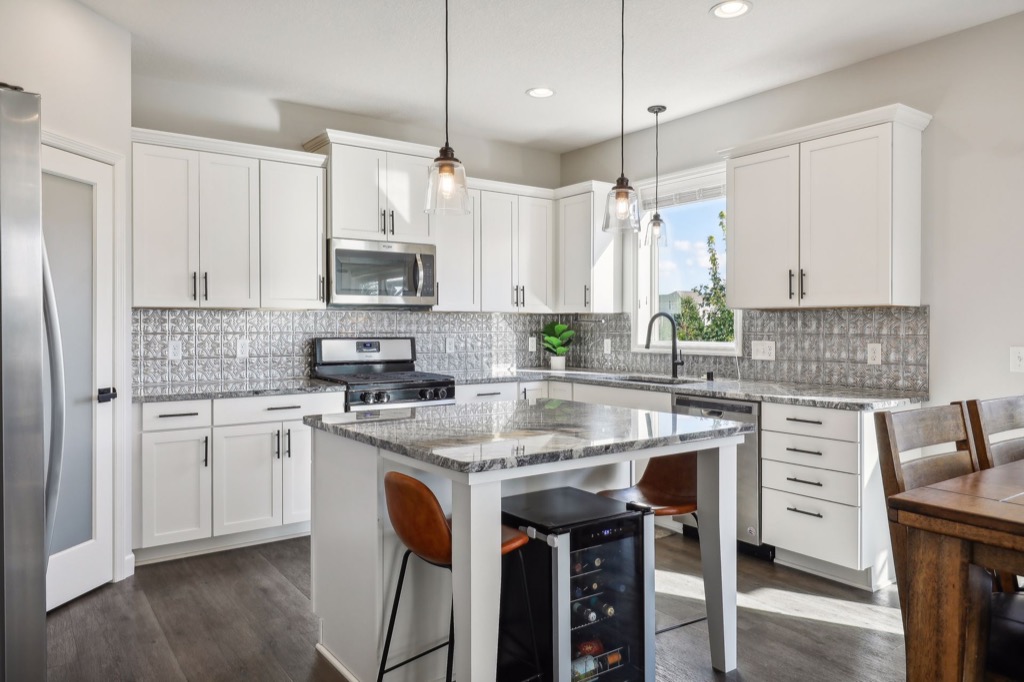 Modern kitchen at 15668 Fairchild Way with white cabinets, stainless steel appliances, a gray tile backsplash, granite countertops, a center island with two stools and a wine fridge, pendant lights, and a dining table with wooden chairs. See PHOTOS for details.