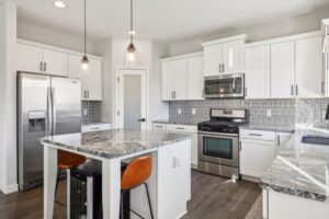 Modern kitchen on Fairchild Way with white cabinets, stainless steel appliances, a gray patterned backsplash, marble countertops, a kitchen island with two orange chairs, pendant lights, and stunning PHOTOS that showcase every detail.