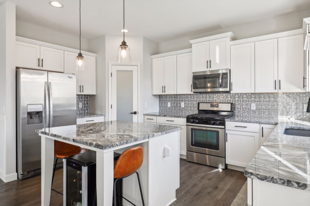 Modern kitchen on Fairchild Way with white cabinets, stainless steel appliances, a gray patterned backsplash, marble countertops, a kitchen island with two orange chairs, pendant lights, and stunning PHOTOS that showcase every detail.