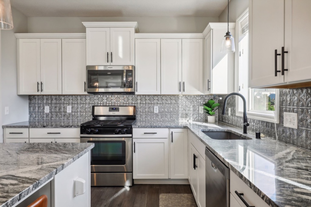 Modern kitchen on Fairchild Way with white cabinets, stainless steel appliances, gray patterned backsplash, marble countertops, black faucet, and pendant lighting. See PHOTOS of a small potted plant by the window above the sink.