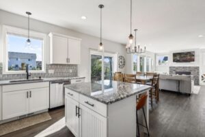 Modern open-concept kitchen and living area at 15668 Fairchild Way with white cabinets, granite island, stainless steel appliances, dining table, and large windows. Living room features a fireplace, mounted TV—see PHOTOS for a closer look.