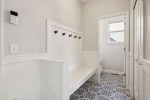 A hallway on Fairchild Way with patterned tile flooring, white paneled walls, a built-in white bench with hooks above it, white doors, and a keypad near the entrance. Sunlight streams in from a window—perfect for PHOTOS.