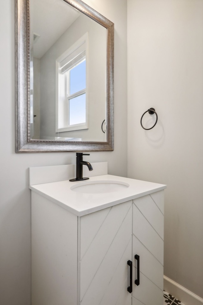 A small modern bathroom on Fairchild Way features a white vanity, black faucet, black cabinet handles, a round black towel ring, a large framed mirror, and a window with daylight streaming in—perfect for stunning PHOTOS.