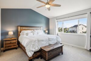 A modern bedroom on Fairchild Way featuring a wooden bed, white bedding, matching nightstands with lamps, a wooden bench at the foot, gray accent wall, and a large window with white curtains and neighborhood views. See PHOTOS for details.