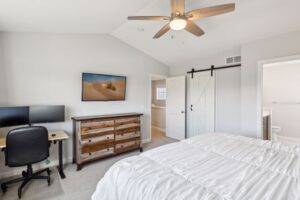 Bright bedroom on Fairchild Way with white bedding, rustic wooden dresser, wall-mounted TV, small desk with dual monitors and chair, ceiling fan, and a sliding barn door leading to a bathroom. View PHOTOS for more details. 15668.