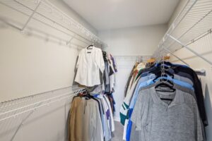 A walk-in closet with wire shelves, neatly organized with hanging clothing including shirts, blouses, jackets, and pants. Light-colored walls enhance the clean look—see PHOTOS for more details of this Fairchild Way space.