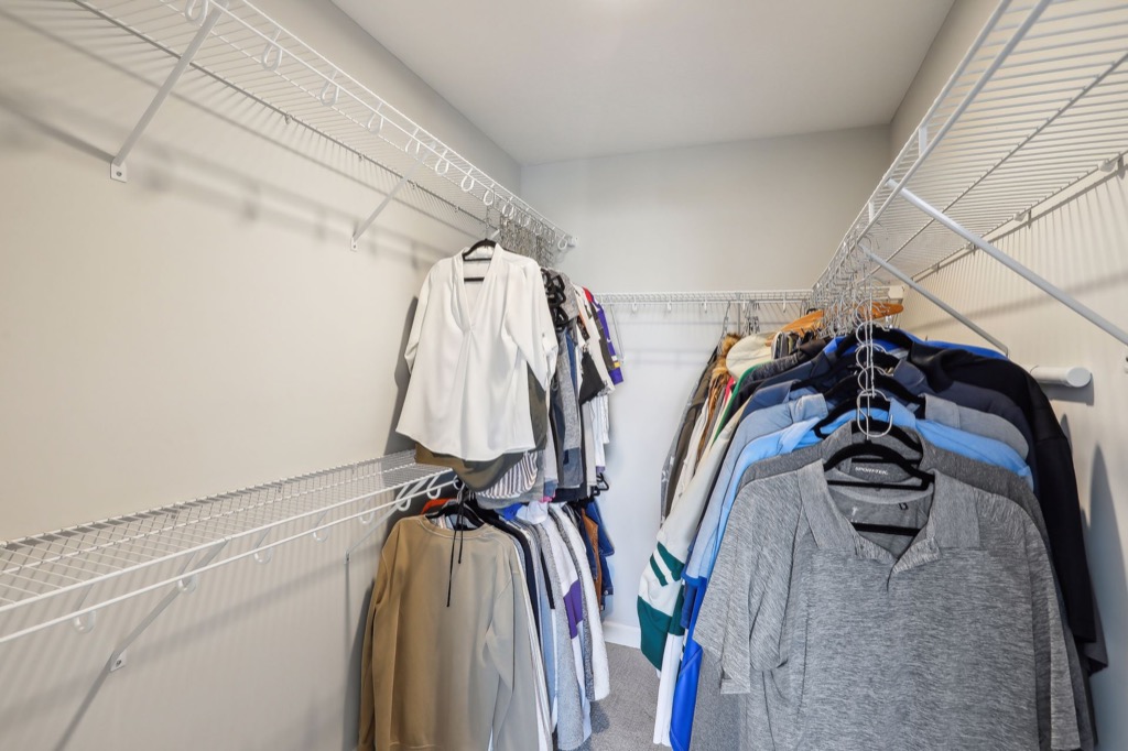 A walk-in closet with wire shelves, neatly organized with hanging clothing including shirts, blouses, jackets, and pants. Light-colored walls enhance the clean look—see PHOTOS for more details of this Fairchild Way space.