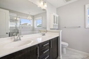 Modern bathroom at 15668 Fairchild Way with a double-sink vanity, dark cabinets, large mirror, white countertop, toilet, wall cabinet, towel rack, and a shower with glass doors next to windows. See photos for details.