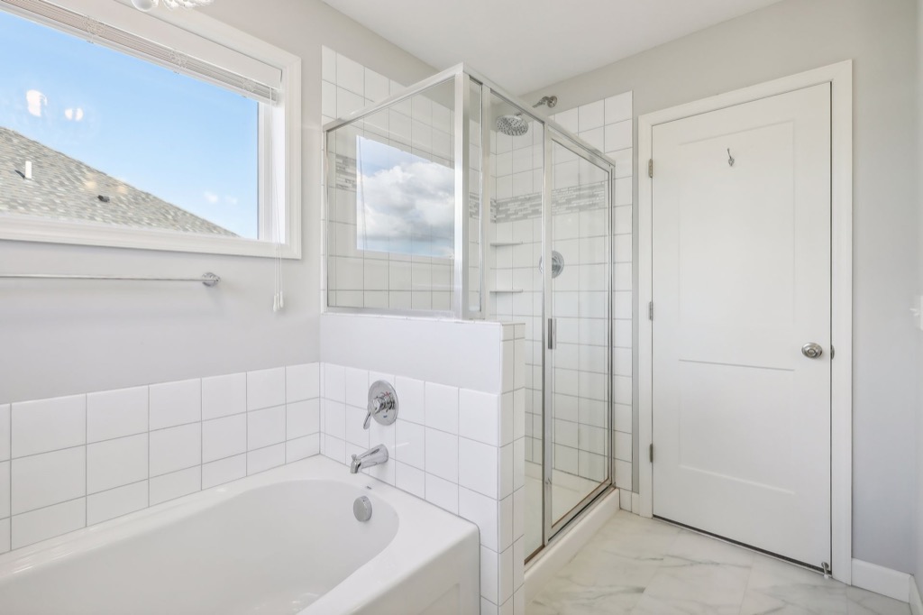 A bright Fairchild Way bathroom with a white bathtub, glass-enclosed shower, tiled walls, marble-patterned floor, and sunlight streaming through the window—see more in photos from 15668.