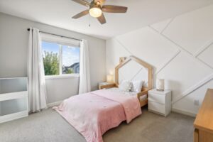 A bright bedroom at 15668 Fairchild Way features a single bed with a pink blanket, white pillows, two white nightstands with lamps, a window with white curtains, beige carpet, and a ceiling fan with wooden blades; see PHOTOS for details.