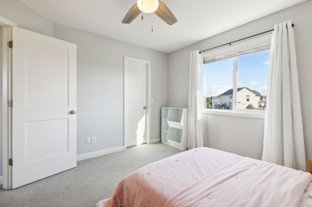 A bright bedroom at 15668 Fairchild Way features light gray walls, carpeted floor, a ceiling fan, two white doors, a window with white curtains, a small shelf, and a bed covered with a pink blanket—perfect for cozy photos.