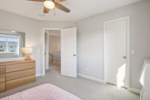 A bright Fairchild Way bedroom with beige carpet, light gray walls, a bed with a pink polka dot blanket, a wooden dresser with lamp, ceiling fan, and two white doors. PHOTOS show natural light streaming in through the window.