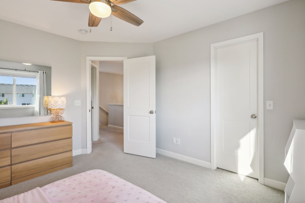 A bright Fairchild Way bedroom with beige carpet, light gray walls, a bed with a pink polka dot blanket, a wooden dresser with lamp, ceiling fan, and two white doors. PHOTOS show natural light streaming in through the window.