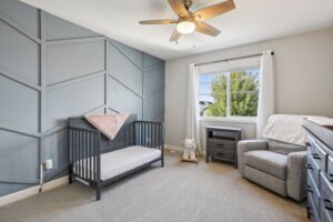 A modern nursery on Fairchild Way features a gray accent wall, black crib, gray armchair, small desk, white-curtained window, plush rocking lamb on beige carpet, and ceiling fan with lights. View more details in the PHOTOS.