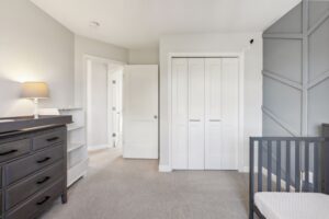 Modern, neutral-toned nursery at 15668 Fairchild Way features a dark dresser, white shelving, gray crib, beige carpet, and white closet doors. A lamp sits on the dresser next to a geometric accent wall. See PHOTOS for more details.