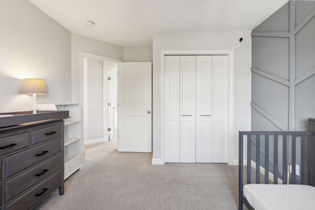 Modern, neutral-toned nursery at 15668 Fairchild Way features a dark dresser, white shelving, gray crib, beige carpet, and white closet doors. A lamp sits on the dresser next to a geometric accent wall. See PHOTOS for more details.