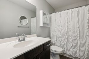 A modern bathroom on Fairchild Way features a dark wood vanity, white countertop, oval sink, round mirror, wall cabinet, toilet, and a shower with a ruffled white curtain. The light gray walls create a fresh look—perfect for PHOTOS.