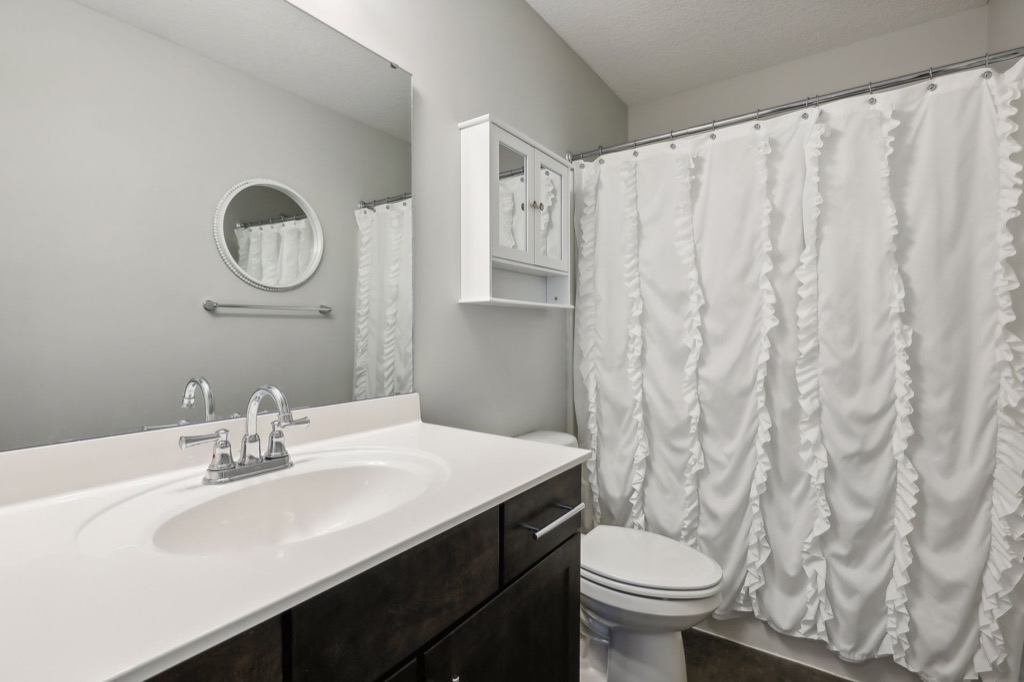 A modern bathroom on Fairchild Way features a dark wood vanity, white countertop, oval sink, round mirror, wall cabinet, toilet, and a shower with a ruffled white curtain. The light gray walls create a fresh look—perfect for PHOTOS.