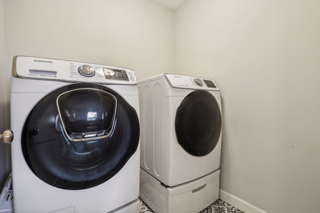 A laundry room on Fairchild Way, 15668, features a front-loading Samsung washer and dryer set, both white with black doors, placed side by side against a light-colored wall on a patterned tile floor—perfect for your photos.