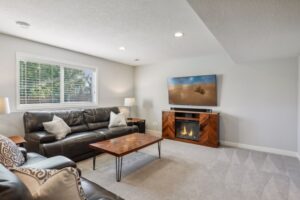 A bright living room on Fairchild Way features a black leather sofa and armchair, wooden coffee table, TV above an electric fireplace, neutral walls, carpeted floor, and a large window with blinds letting in natural light. See PHOTOS.