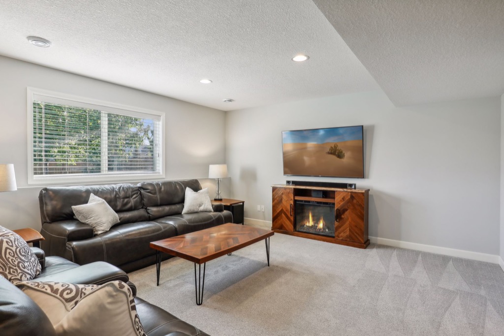 A bright living room on Fairchild Way features a black leather sofa and armchair, wooden coffee table, TV above an electric fireplace, neutral walls, carpeted floor, and a large window with blinds letting in natural light. See PHOTOS.