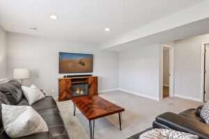 A modern living room on Fairchild Way features a black leather sofa, patterned pillows, a wooden coffee table, and an electric fireplace beneath a TV. PHOTOS reveal light gray walls, carpeted floor, and two doorways on the right.