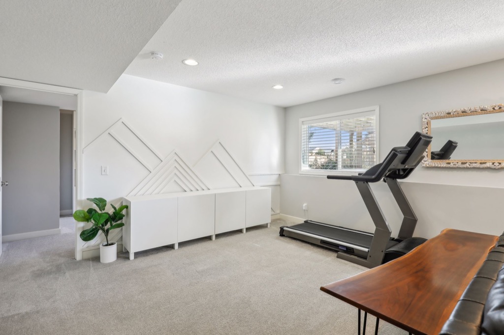 A bright home gym room on Fairchild Way with a treadmill facing a window, a modern white geometric accent wall, beige carpet, a potted plant, and decorative mirror above a wooden table—see PHOTOS for more details.