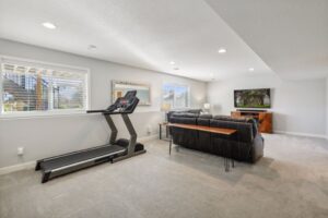 A bright, modern basement room on Fairchild Way features a treadmill near two windows, a large black sectional sofa, a wood TV console, and a flat-screen TV. The carpeted floor and recessed lighting create a cozy atmosphere—see PHOTOS for details.