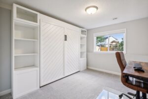 Bright home office at 15668 Fairchild Way with a large window, wooden desk, brown chair, and a white built-in shelving unit featuring chevron-patterned doors. PHOTOS reveal light gray carpet and neutral walls for a clean, modern look.