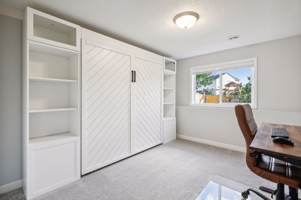 Bright home office at 15668 Fairchild Way with a large window, wooden desk, brown chair, and a white built-in shelving unit featuring chevron-patterned doors. PHOTOS reveal light gray carpet and neutral walls for a clean, modern look.