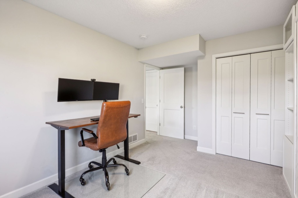 A minimalist home office on Fairchild Way features a brown leather chair at a wooden desk, a large monitor, and keyboard. Light gray walls, carpeted floor, and double-door closet complete the space—see more in the PHOTOS.