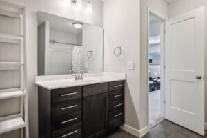 Modern bathroom at 15668 Fairchild Way features a dark wood vanity, large mirror, white countertop, built-in shelves on the left, towel ring, and an open door revealing another room with a couch. See PHOTOS for details.