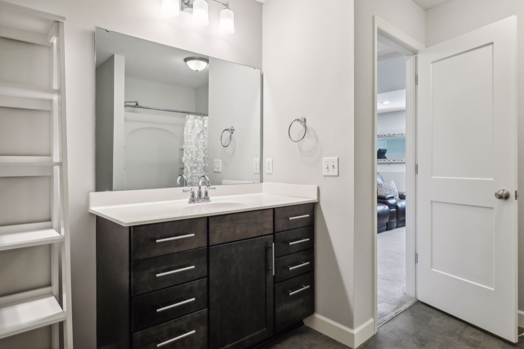 Modern bathroom at 15668 Fairchild Way features a dark wood vanity, large mirror, white countertop, built-in shelves on the left, towel ring, and an open door revealing another room with a couch. See PHOTOS for details.