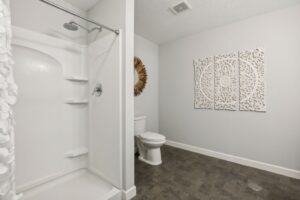A modern bathroom at 15668 Fairchild Way features a white walk-in shower, toilet, decorative wall mirror, and intricate white wall art above dark flooring. The light gray walls and minimalistic design create a clean look. PHOTOS highlight its stunning details.