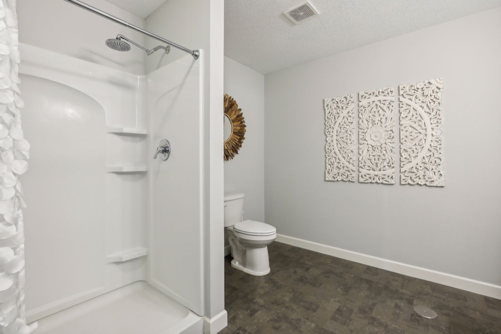 A modern bathroom at 15668 Fairchild Way features a white walk-in shower, toilet, decorative wall mirror, and intricate white wall art above dark flooring. The light gray walls and minimalistic design create a clean look. PHOTOS highlight its stunning details.