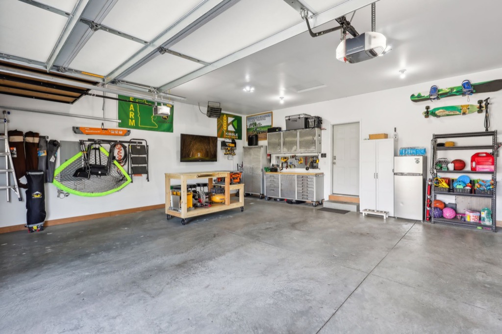 A clean, organized garage at 15668 Fairchild Way features tools, sports equipment, and storage shelves along white walls. A workbench stands in the middle, and the open garage door lets in natural light for perfect PHOTOS.