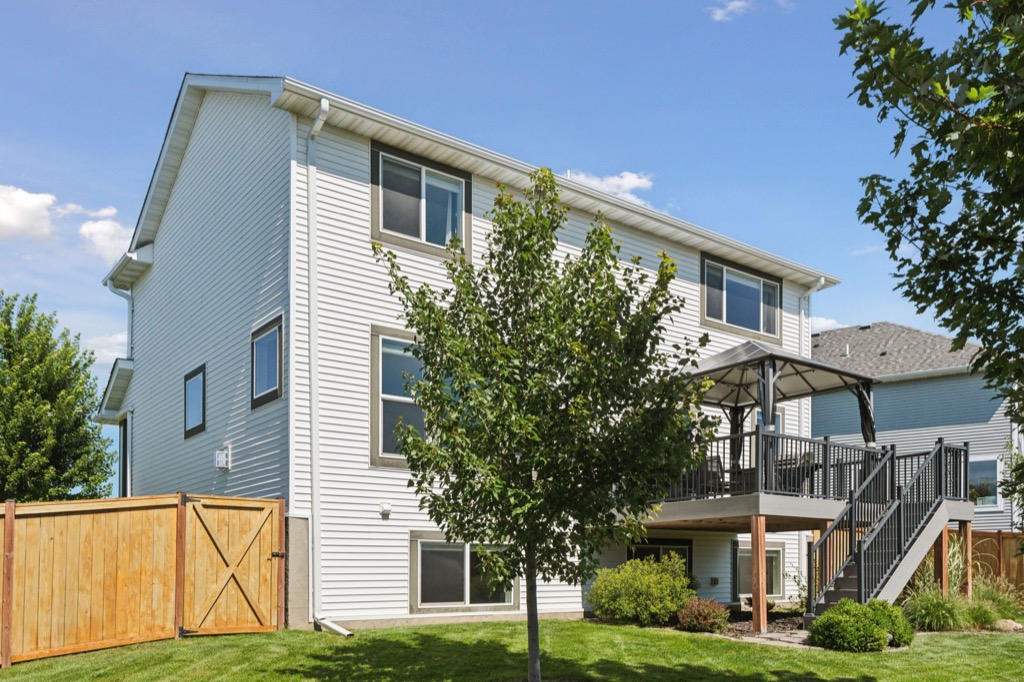 A two-story house on Fairchild Way with light-colored siding, large windows, a raised deck with stairs, a wooden fenced yard, green grass, trees, and shrubs under a clear blue sky—see photos 15668 for more details.