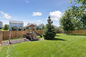 A spacious backyard on Fairchild Way with green grass, a wooden playset with swings and slide, a grand evergreen tree, and a wood fence—perfect for real estate photos. Several houses and trees sit in the background beneath a blue sky with clouds.
