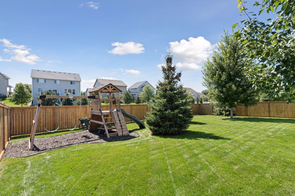 A spacious backyard on Fairchild Way with green grass, a wooden playset with swings and slide, a grand evergreen tree, and a wood fence—perfect for real estate photos. Several houses and trees sit in the background beneath a blue sky with clouds.