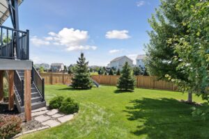 A sunny backyard on Fairchild Way with green grass, several trees, a wooden fence, houses in the background, and a staircase leading down from a deck on the left side. The sky is blue with scattered clouds—perfect for photos.